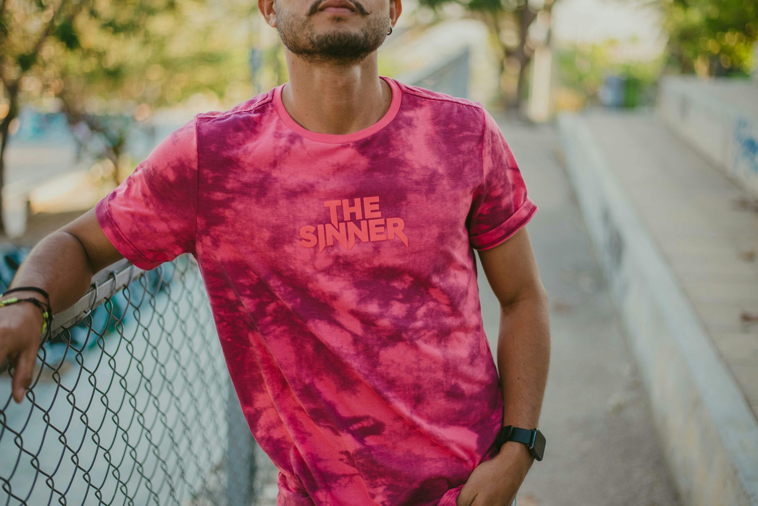 Young man in pink tie-dye t-shirt posing casually by a chain-link fence outdoors.